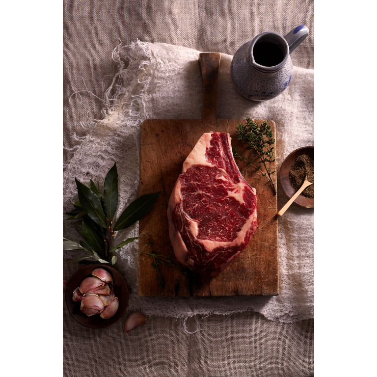 A raw marbled rib eye steak on a chopping board with garnishes decorating the table